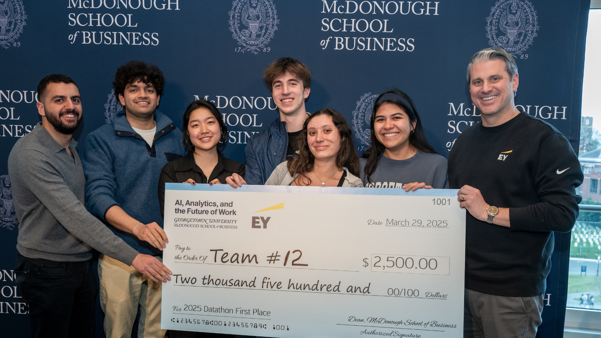 A group of six McDonough business students and an EY staff member stand in front of a McDonough School of Business backdrop holding a large check for $2,500